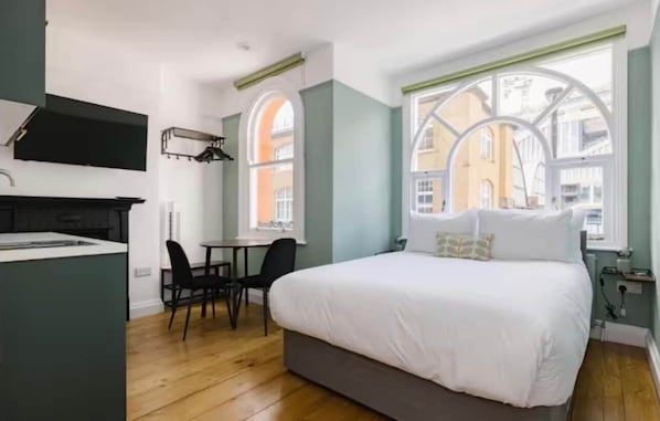 A compact hotel room with a bed, a small kitchenette, a dining area, and large windows offering a view of the city.