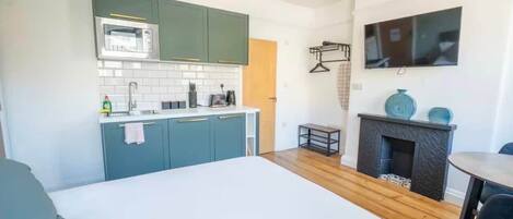A compact kitchen with a microwave, sink, and green cabinets. There's a small table and a fireplace with decorative items.