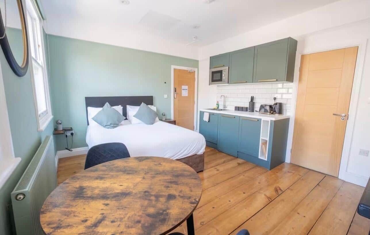 A compact room with a bed, a small table, a kitchenette, and a door.