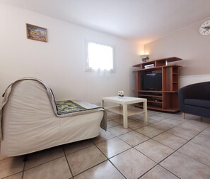 Una habitación pequeña y luminosa con una cama individual, un televisor, una mesita y una silla.