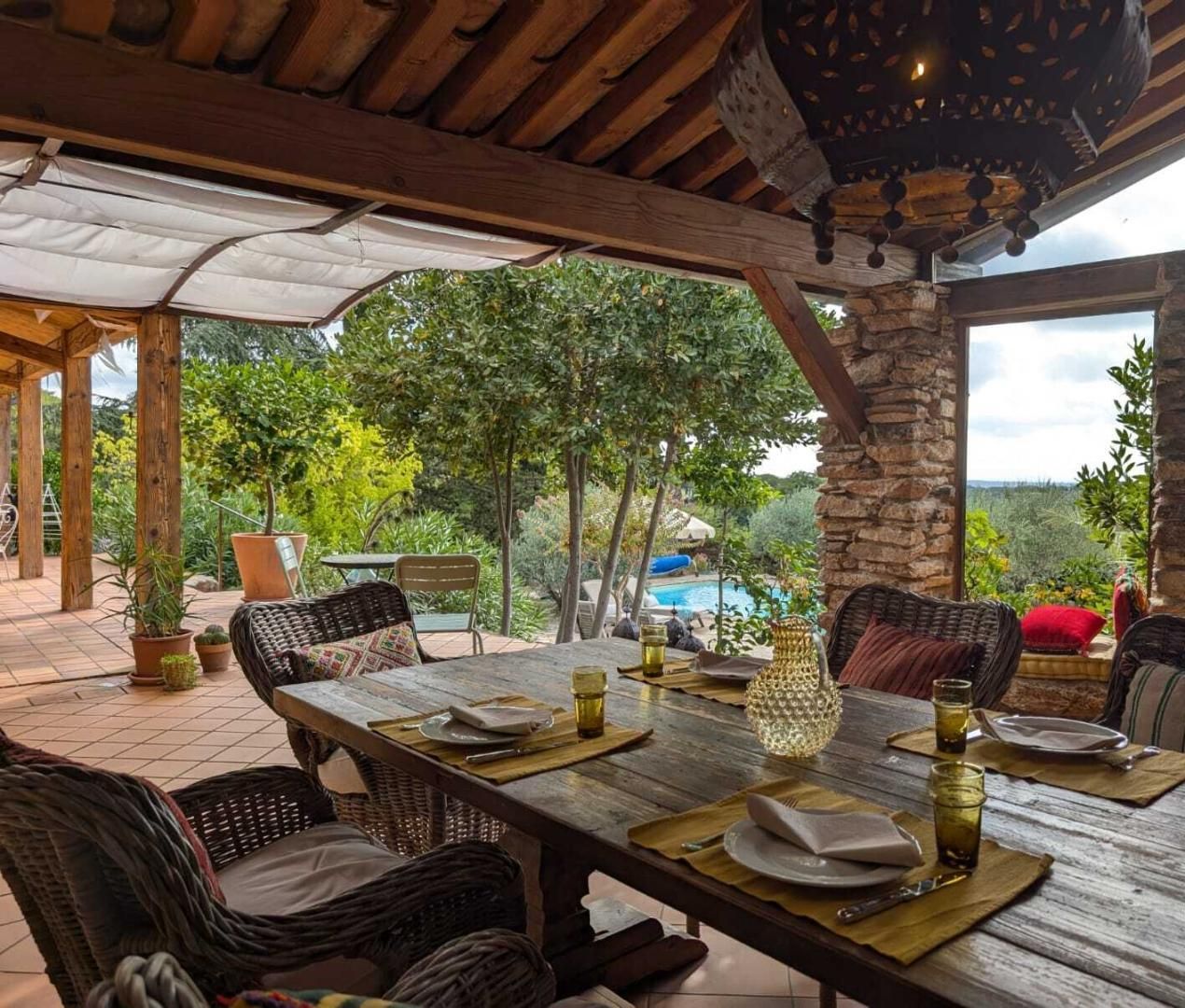 Un área de comedor al aire libre con una mesa de madera dispuesta para dos personas, rodeada de sillas de mimbre, un pilar de piedra y vistas a una piscina y árboles al fondo.