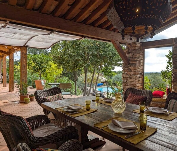 An outdoor dining area with a wooden table set for two, surrounded by wicker chairs, with a stone pillar and a view of a swimming pool and trees in the background.