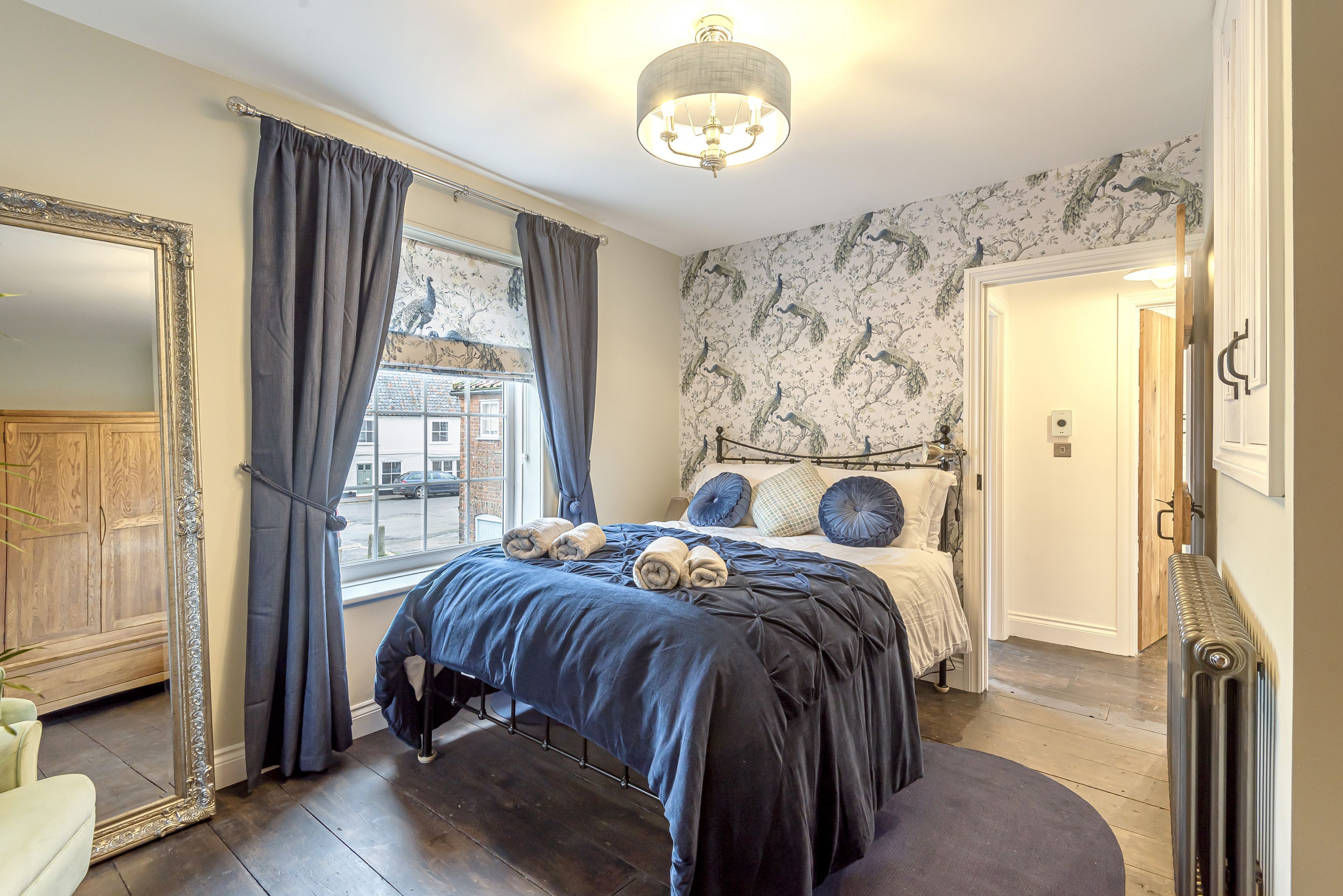 A bedroom with a large bed, dark curtains, a mirror, and a window with a view of buildings outside.