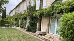 Un bâtiment historique doté d’une façade en pierre, de volets blancs et d’un jardin bien entretenu avec une pelouse et une table entourée de chaises.