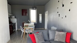 Un salon moderne avec un canapé gris, des coussins rouges et gris, un coin repas avec une table et des chaises blanches, et une cuisine en arrière-plan.