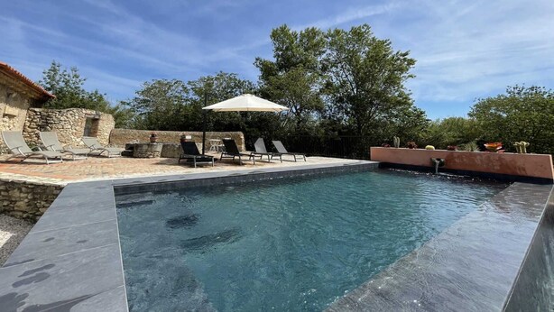 Une piscine entourée de murs de pierre et d’arbres, avec des chaises longues et un parasol.