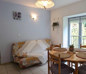 Uma sala de estar aconchegante com um sofá, uma mesa de jantar com jogos americanos, uma planta pendente e uma janela com cortinas de renda.