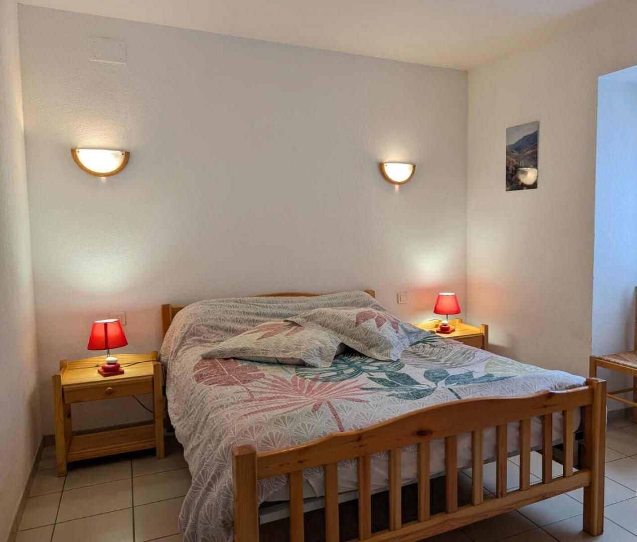 A bedroom with a wooden bed, two bedside lamps, a window with lace curtains, and a small chair.