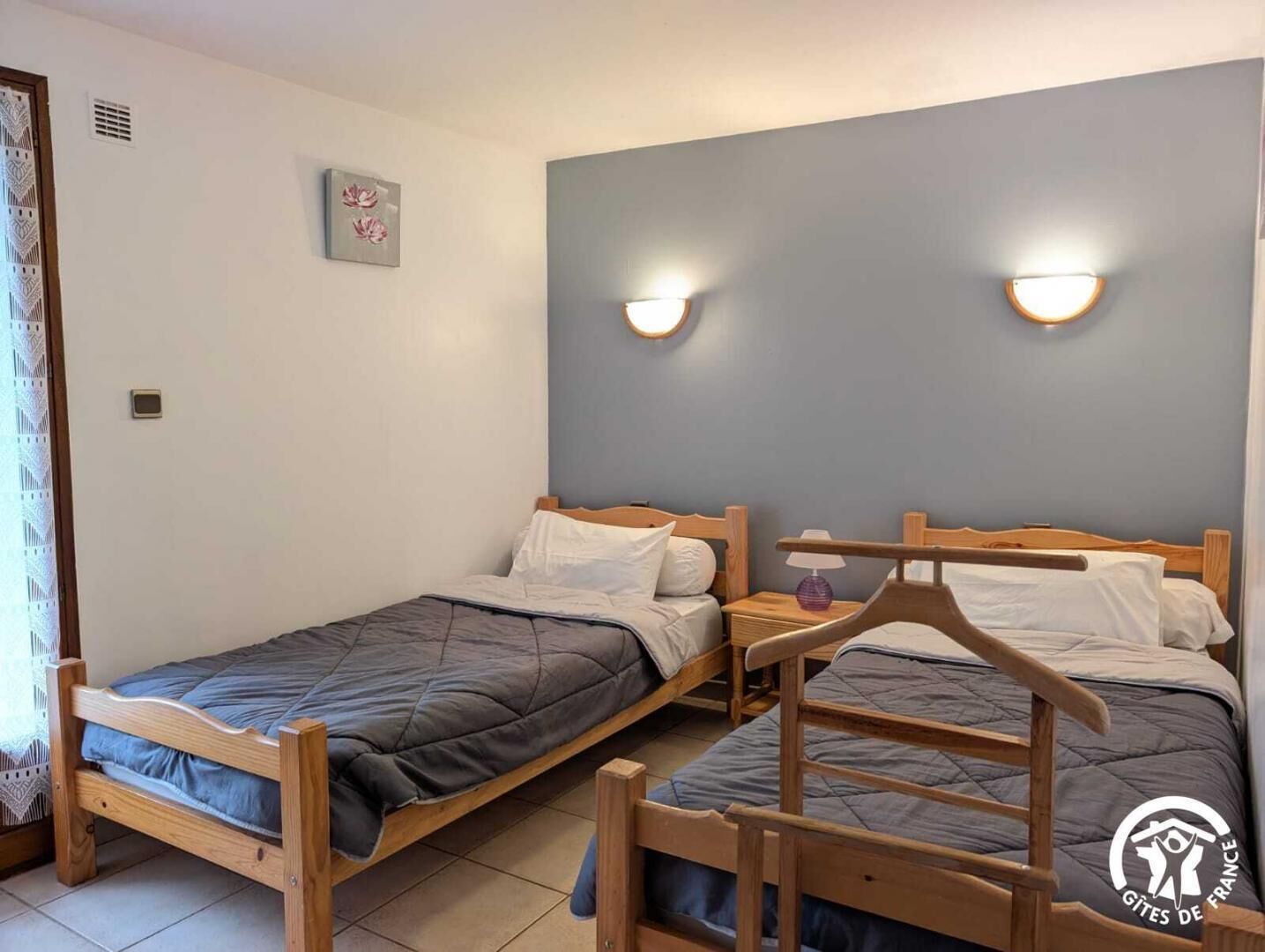 Ein Zimmer mit zwei hölzernen Betten, jeweils mit einer grauen Bettdecke und weißen Kissen. An der Wand sind Leuchten befestigt und ein kleines Bild hängt dort.
