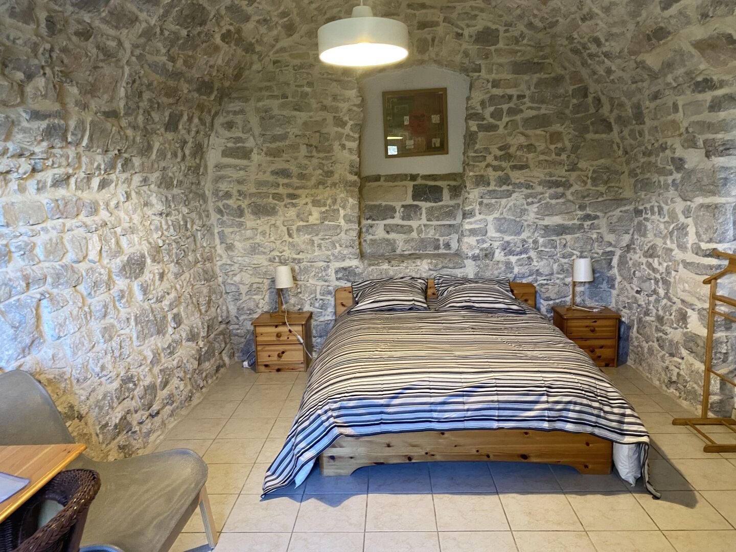 Habitación con paredes de piedra, una cama, dos mesitas de noche, una silla y una lámpara colgante.