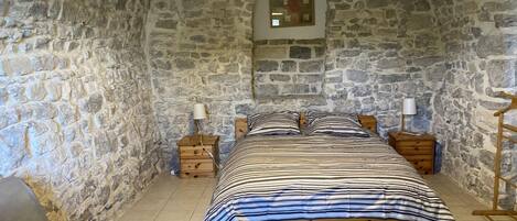 Habitación con paredes de piedra, una cama, dos mesitas de noche, una silla y una lámpara colgante.