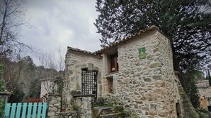 Ein Steinhaus mit einem kleinen Fenster und einer Tür, umgeben von Bäumen und einem blauen Zaun.