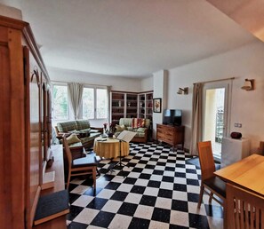A room with a checkered floor, a wooden dining table, a sofa, a television, and a bookshelf.