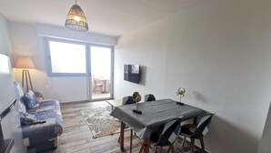 A modern living room with a sofa, dining table, and chairs. There is a television mounted on the wall, a lamp, and a patterned rug on the floor.