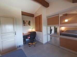 A bedroom with a bed, a desk with a chair, a painting on the wall, and a sliding door leading to another room. - Bergerie du Pic Saint-Loup (SAINT-ANDRE-DE-BUEGES)