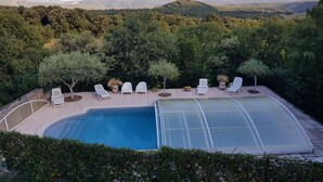 A swimming pool with a glass enclosure, surrounded by white lounge chairs and small trees, set against a backdrop of hills and a clear sky.