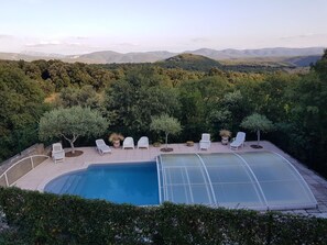 A swimming pool with a glass enclosure, surrounded by white lounge chairs and small trees, set against a backdrop of hills and a clear sky.