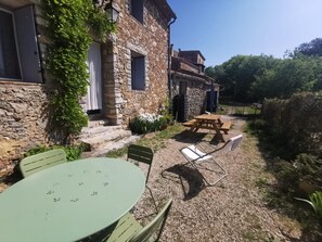 A stone building with a patio area featuring a table and chairs, a wooden bench, and a small garden with plants.