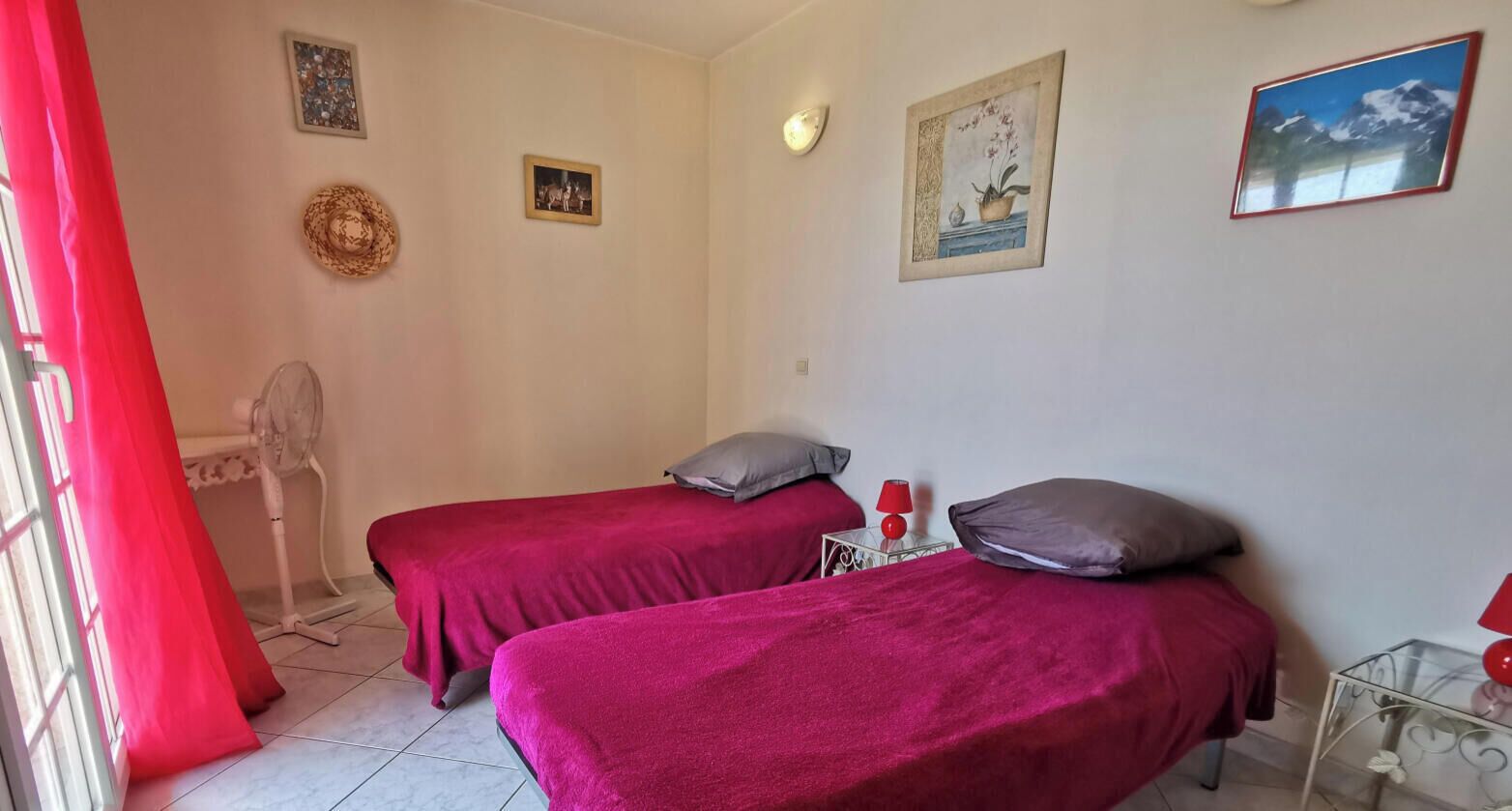 Ein Zimmer mit zwei Betten, einem Ventilator und einem Fenster mit roten Vorhängen.
