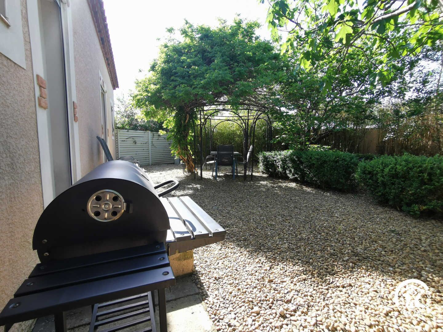 A barbecue grill in a backyard with a gravel path, a wooden bench, and a pergola.