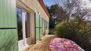 Una casa con contraventanas verdes, un patio con baldosas y una mesa con mantel transparente.