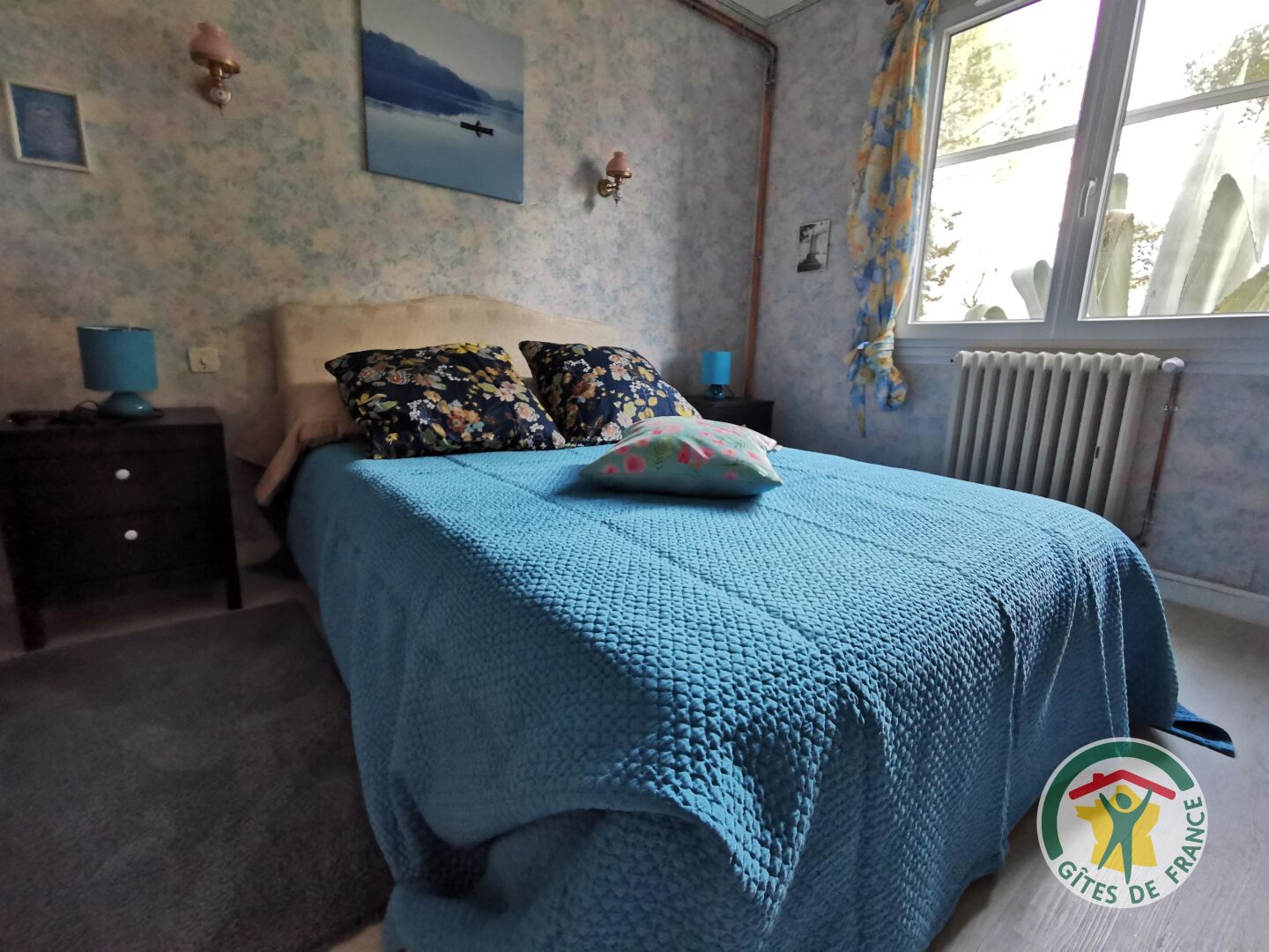 A bedroom with a blue bedspread, floral pillows, a nightstand with a lamp, and a window with a view of trees.