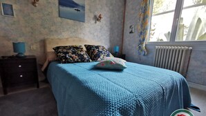 Un dormitorio con colcha azul, almohadas estampadas, una mesita de noche con lámpara y ventana con vistas a árboles.