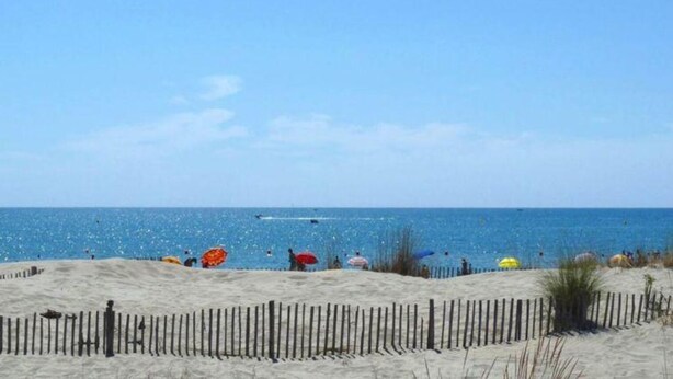 Ein sandiger Strand mit einem Holzzaun im Vordergrund und blauem, wolkenlosem Himmel.