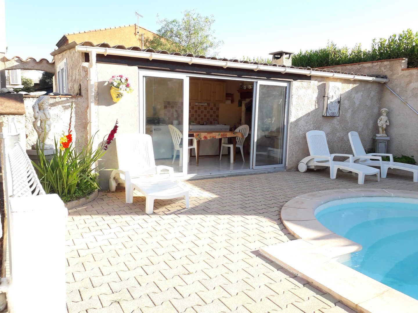 A patio area with a pool, white lounge chairs, a dining table with chairs, and a sliding glass door leading to a kitchen.