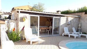 Eine Terrassenanlage mit Pool, weißen Liegestühlen, einem Esstisch mit Stühlen und einer Glastür zum Küchenbereich.