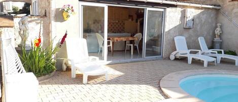 A patio area with a pool, white lounge chairs, a dining table with chairs, and a sliding glass door leading to a kitchen.
