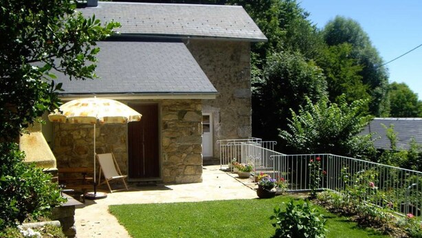 A stone house with a grey roof, a small patio with a white railing, and a garden with red flowers.