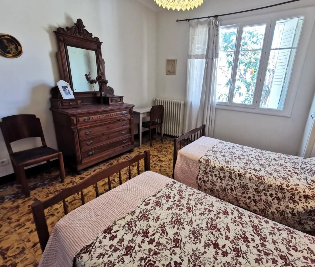 A room with two beds, a wooden dresser, a chair, a desk, and a window with curtains.
