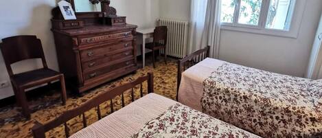 A room with two beds, a wooden dresser, a chair, a desk, and a window with curtains.