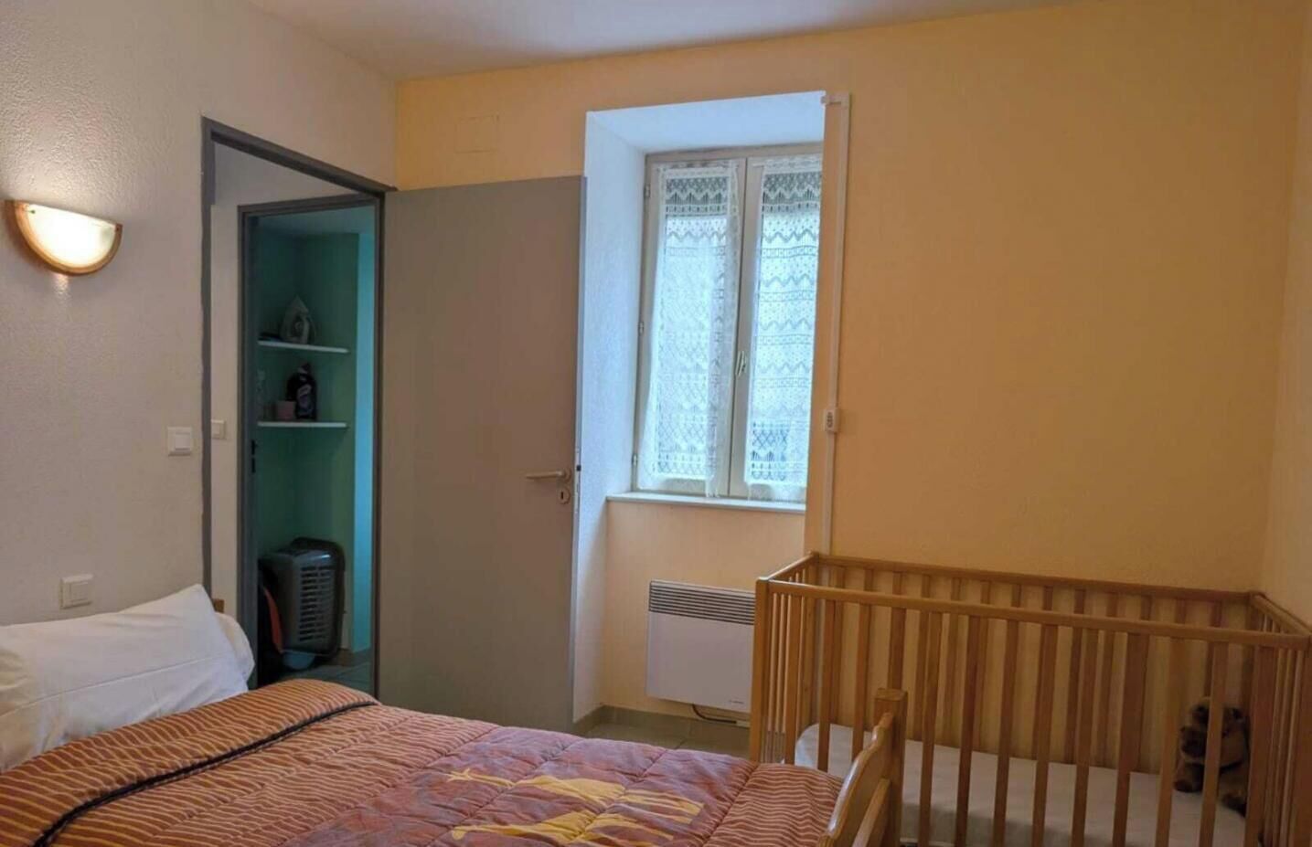 Une chambre à coucher avec un lit, un berceau, une armoire et une fenêtre ornée de rideaux en dentelle.