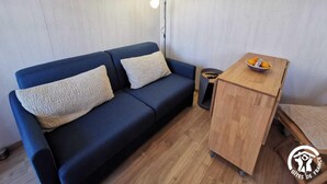Ein gemütlicher Raum mit einem dunkelblauen Sofa, zwei hellen Kissen, einem kleinen Holztisch mit Obstschale und einer Stehlampe.