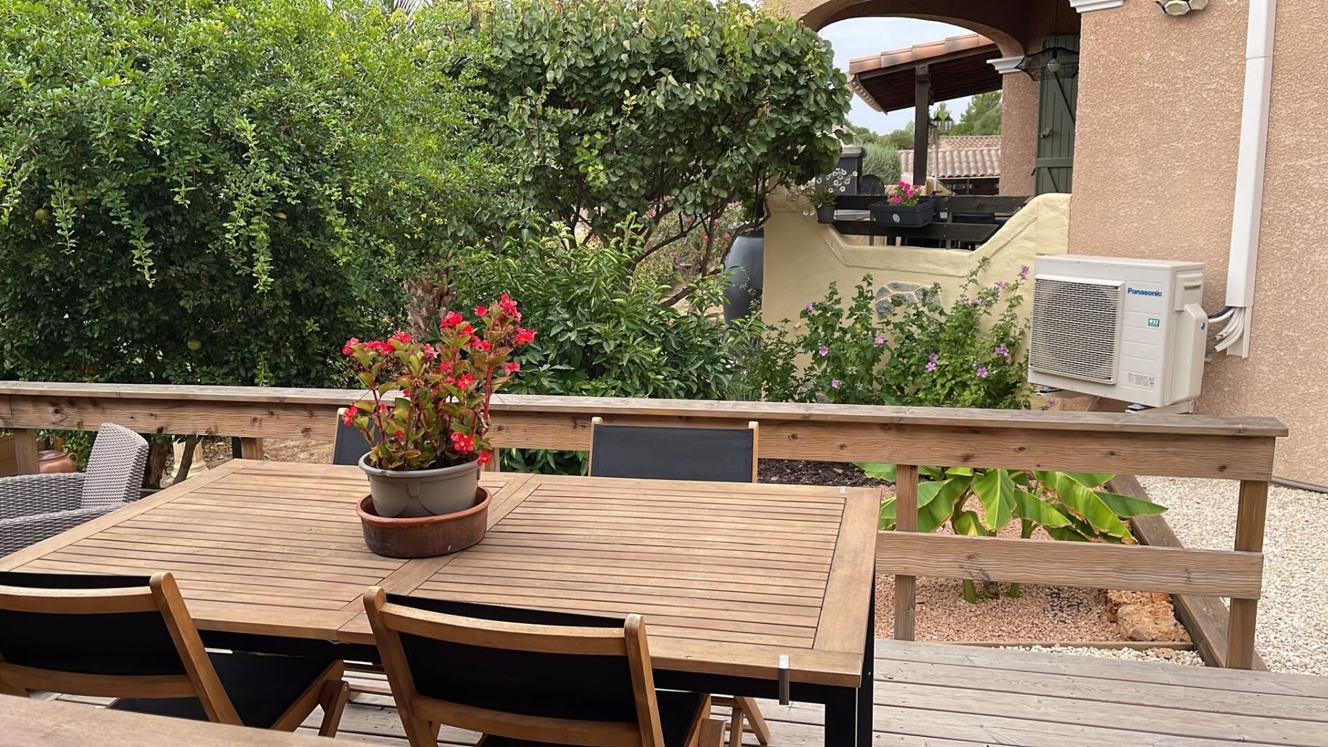 Eine Außenpatio mit einem Holztisch und Stühlen, einem Blumentopf mit roten Blüten und einer Panasonic Klimaanlage.