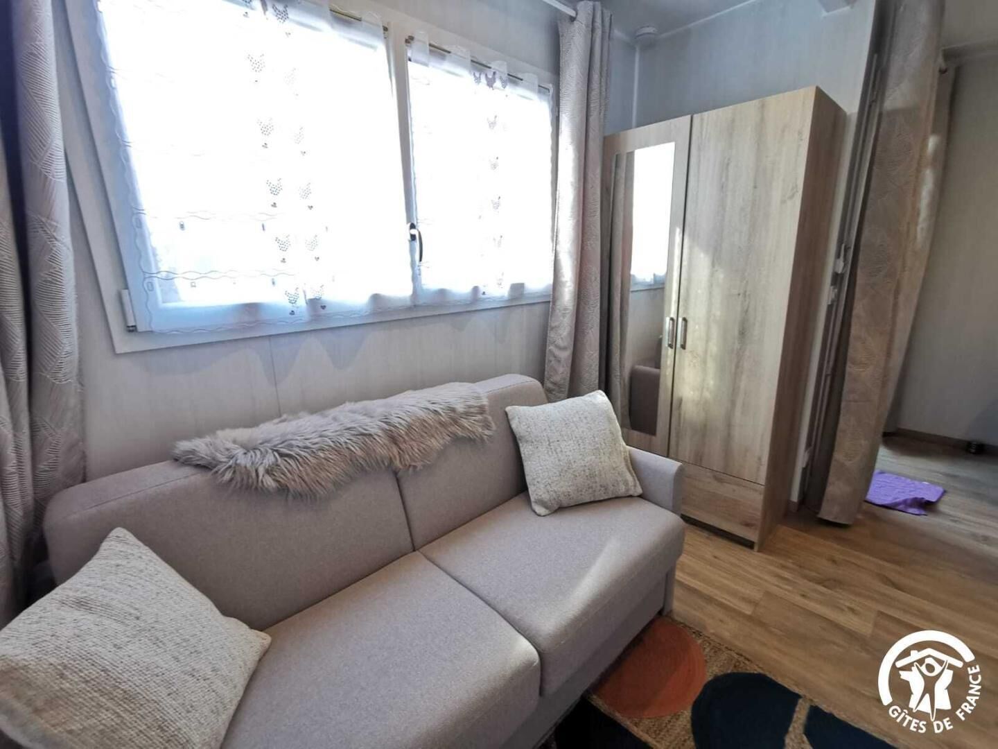 A neutral-toned sofa with two cushions, a window with lace curtains, and a wooden wardrobe.