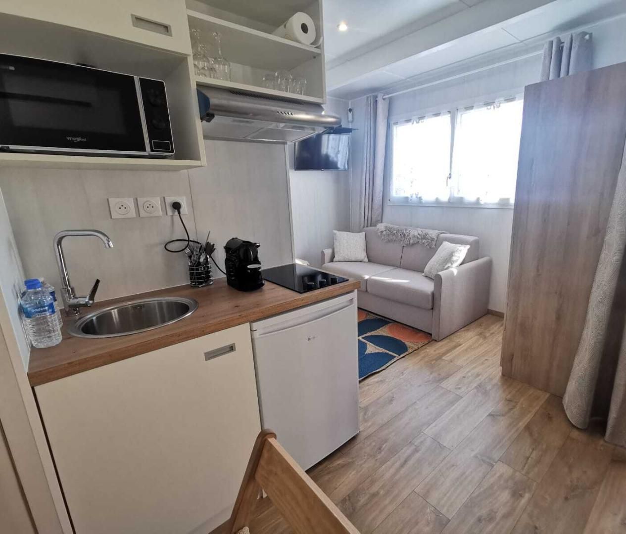 Eine kompakte Küche mit Mikrowelle, Spüle und Geschirrspüler. Im Wohnbereich befindet sich eine Couch und ein Fenster mit Vorhängen.