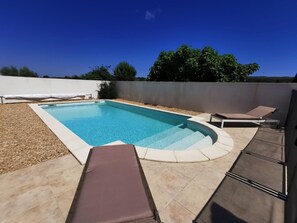 A rectangular swimming pool with a tiled deck, surrounded by a low wall and a few trees in the background.