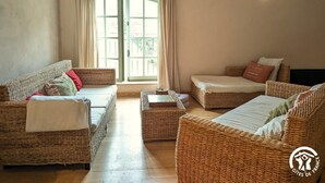 Een ruime kamer met rieten meubels, een groot raam met gordijnen en wandlampen.