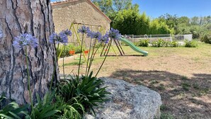 A playground with swings and a slide, surrounded by trees and a stone building. - Côté Cour - MARAUSSAN - Gîte (MARAUSSAN)