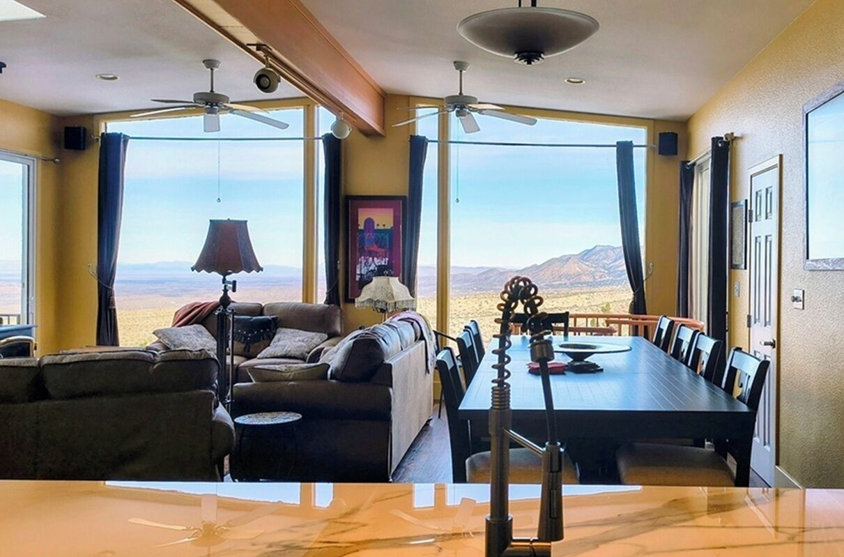 A room with a sofa, dining table, chairs, ceiling fans, and large windows offering a view of the mountains.