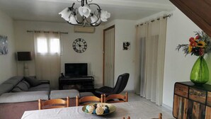A living room with a sofa, a TV, a dining table with chairs, a chandelier, and a cabinet with a vase of flowers.