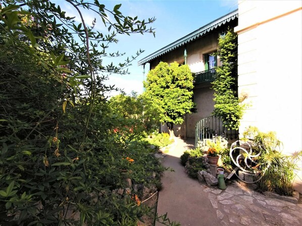 Un sendero conduce a un edificio de estilo arquitectónico tradicional, rodeado de abundante vegetación y plantas en macetas.