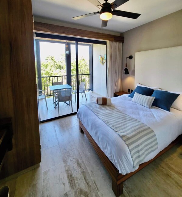 A hotel room with a large bed, a ceiling fan, a balcony with a table and chairs, and a view of greenery outside.