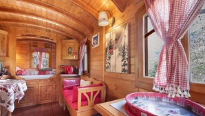 A cozy cabin interior with wooden walls, a bed with red bedding, a dining table, and a window with a view of trees.
