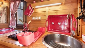 A wooden cabin interior with a kitchen area, including a sink, red dish rack, and wooden shelves with red mugs.