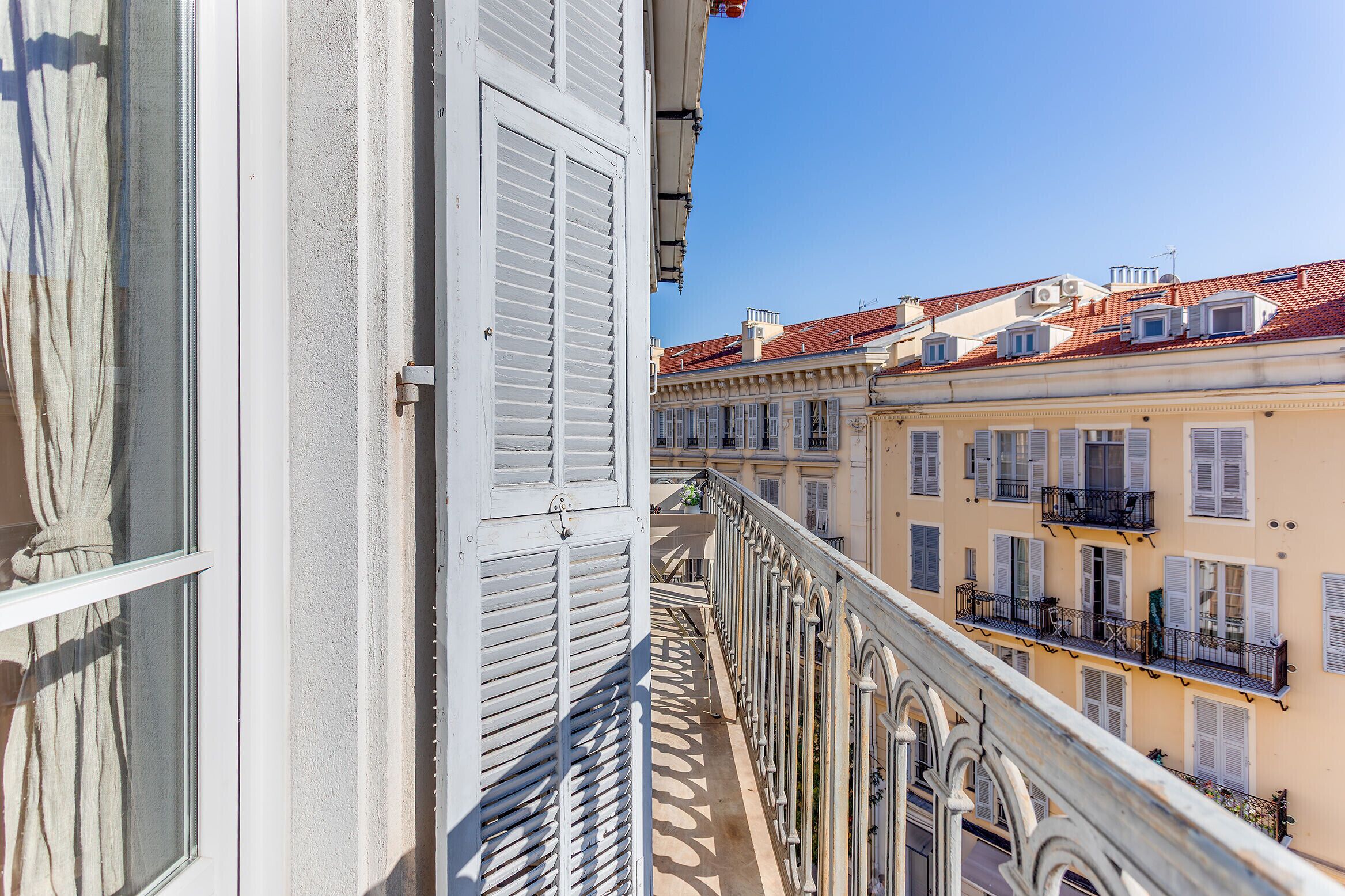 A balcony with white shutters, a view of adjacent buildings with red-tiled roofs, and a clear blue sky.