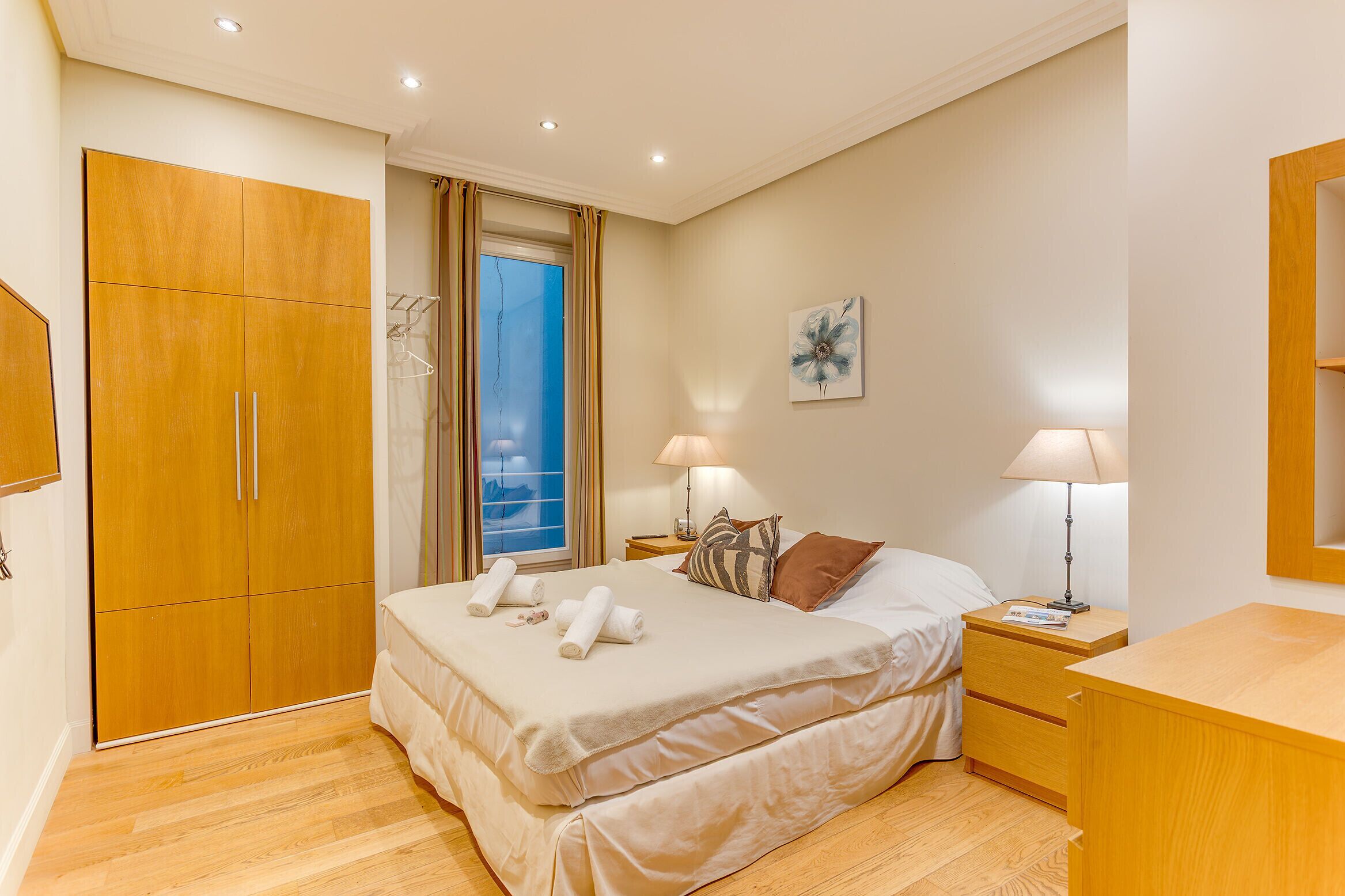 A bedroom with a large wardrobe, a bed with two pillows, a nightstand with a lamp, and a window with curtains.